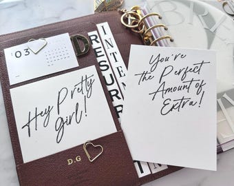 Może przedstawiać: Brązowy skórzany planer ze złotymi akcentami, zawierający karty z napisami "Hey Pretty girl!" i "You're the Perfect Amount of Extra!" Widoczny jest również mały kalendarz i spinacze do papieru w kształcie serca.