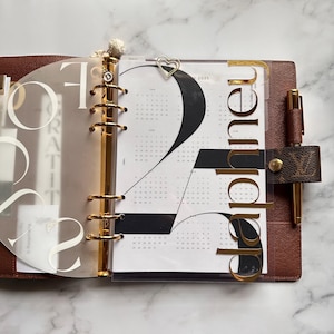 Op de afbeelding: Open bruin leren planner met gouden accenten, inclusief een penhouder en bindringen. De planner is opengeklapt op een kalenderpagina met het nummer 2 in zwart en gouden verdelers die "September" spellen. Een gouden hartvormige paperclip is bevestigd.