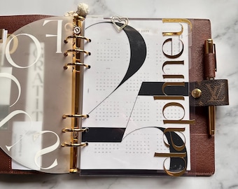 Può includere: Un'agenda ad anelli in pelle marrone con una penna in metallo dorato, una graffetta a forma di cuore e divisori traslucidi. L'agenda mostra una pagina del calendario 2025 con una grande tipografia nera e il nome Daphne in lettere dorate.