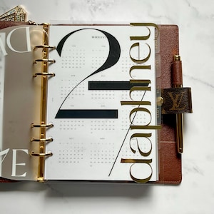 May include: A brown leather ring-bound planner featuring a gold-toned metal mechanism, a gold pen, and a clear plastic dashboard overlay. The dashboard displays the large black text 21 and the gold script text planner over a calendar page.
