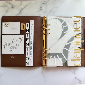 Può includere: Agenda in pelle marrone aperta con dettagli dorati, con inserti con i testi "Hey Pretty Girl!" e "Resurrection". Include una scheda calendario e una sovrapposizione trasparente con le parole "Breathe" e "Clarity" in oro.