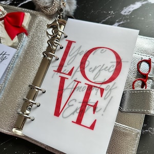 Puede incluir: Carpeta plateada con un inserto transparente con la palabra "LOVE" en rojo, con texto de escritura adicional. Gafas rojas guardadas en un bolsillo. Un lazo rojo y un dije decorativo son visibles.