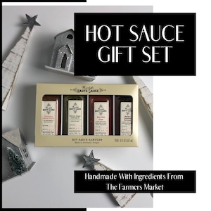 Puede incluir: Un juego de regalo de salsa picante con cuatro botellas diferentes. El texto en la caja dice "Hot Sauce Gift Set" y "Handmade With Ingredients From The Farmers Market". La imagen incluye estrellas decorativas y pequeñas casas.