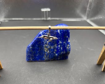 May include: A small, silver-colored whale charm suspended from a wooden dowel. The charm is detailed with fins and a tail, and hangs in front of a dark blue, textured stone. The background is a neutral gray.