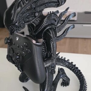 Op de afbeelding: Een zwart 3D-geprint Xenomorph-figuur uit de Alien-franchise, afgebeeld in een knielende houding. Het figuur heeft een lange, gesegmenteerde staart en een gladde, langwerpige kop en houdt een zwarte draadloze gamecontroller in de handen.