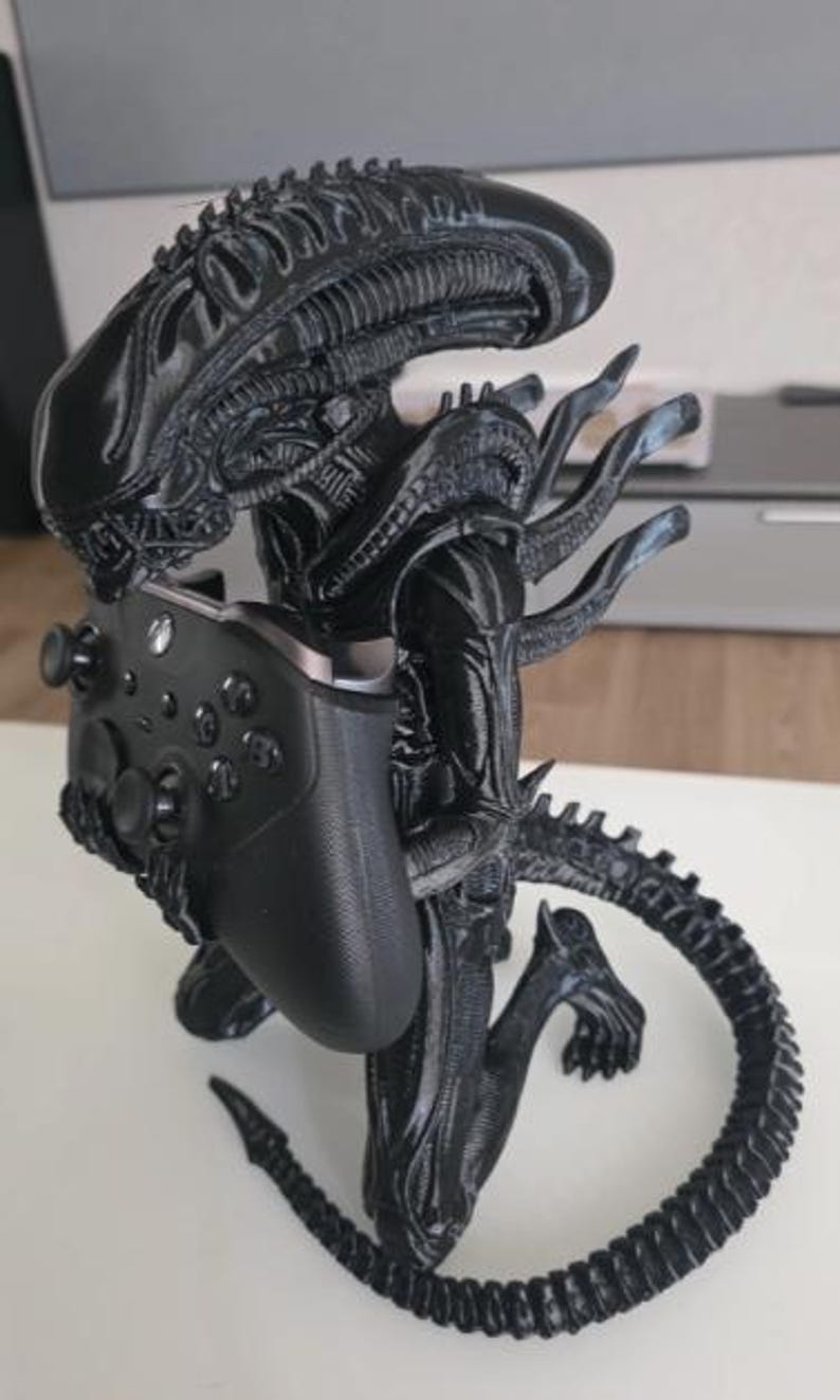 Op de afbeelding: Een zwart 3D-geprint Xenomorph-figuur uit de Alien-franchise, afgebeeld in een knielende houding. Het figuur heeft een lange, gesegmenteerde staart en een gladde, langwerpige kop en houdt een zwarte draadloze gamecontroller in de handen.