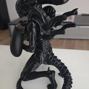 Op de afbeelding: Een zwart 3D-geprint Xenomorph-figuur uit de Alien-franchise, ontworpen als controllerstandaard. Het wezen staat in een gehurkte houding met zijn lange, gesegmenteerde staart opgerold aan de basis en houdt een zwarte gamecontroller op zijn rug.