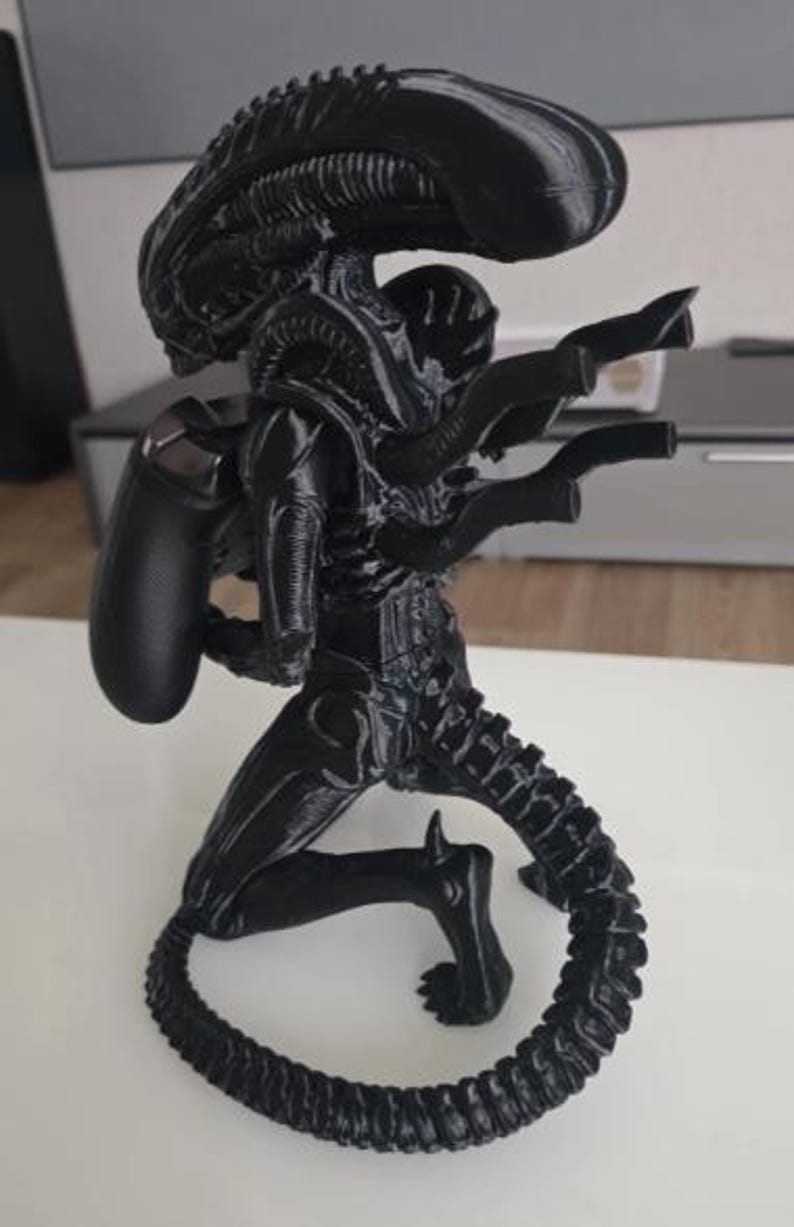 Op de afbeelding: Een zwart 3D-geprint Xenomorph-figuur uit de Alien-franchise, ontworpen als controllerstandaard. Het wezen staat in een gehurkte houding met zijn lange, gesegmenteerde staart opgerold aan de basis en houdt een zwarte gamecontroller op zijn rug.
