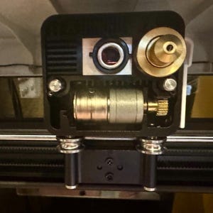 Può includere: Primo piano di un dispositivo meccanico nero e dorato con vari componenti. Il dispositivo presenta una lente, parti metalliche cilindriche e un componente circolare color oro. Il design include viti e un elemento simile a un ingranaggio.