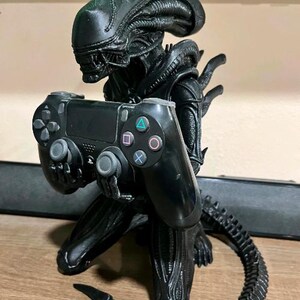 Op de afbeelding: Een zwart Xenomorph-figuurtje in een knielende houding, dat een zwarte PlayStation 4-controller vasthoudt. Het wezen heeft een gladde, langwerpige kop, een gesegmenteerd lichaam en een lange, gebogen staart die langs de basis loopt. De controller heeft standaardknoppen, waaronder de driehoek-, cirkel-, kruis- en vierkantsymbolen.
