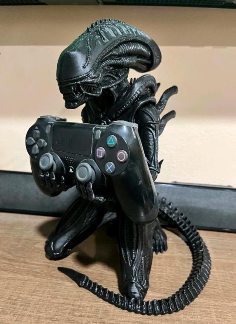 Op de afbeelding: Een zwart Xenomorph-figuurtje in een knielende houding, dat een zwarte PlayStation 4-controller vasthoudt. Het wezen heeft een gladde, langwerpige kop, een gesegmenteerd lichaam en een lange, gebogen staart die langs de basis loopt. De controller heeft standaardknoppen, waaronder de driehoek-, cirkel-, kruis- en vierkantsymbolen.