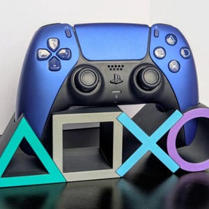 Puede incluir: Un mando de videojuegos azul metálico descansa sobre un soporte negro con forma de símbolos de PlayStation: un triángulo turquesa, un cuadrado gris, una X azul claro y un círculo morado. El mando tiene joysticks negros y botones estándar.