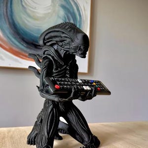 Op de afbeelding: Een matzwart Xenomorph-figuurtje uit de Alien-franchise, afgebeeld in een knielende houding. Het wezen heeft een langwerpig hoofd en getextureerde, biomechanische details. Het houdt een zwarte afstandsbediening voor de televisie vast.