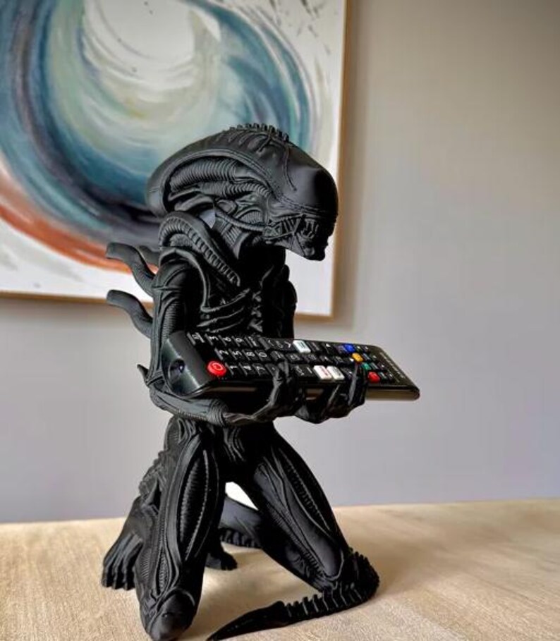 Op de afbeelding: Een matzwart Xenomorph-figuurtje uit de Alien-franchise, afgebeeld in een knielende houding. Het wezen heeft een langwerpig hoofd en getextureerde, biomechanische details. Het houdt een zwarte afstandsbediening voor de televisie vast.