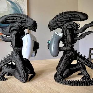 May include: Two black Xenomorph figures in a kneeling position, each holding a video game controller. The figure on the left holds a white controller, while the figure on the right holds a gray controller. Both figures feature detailed ridges.