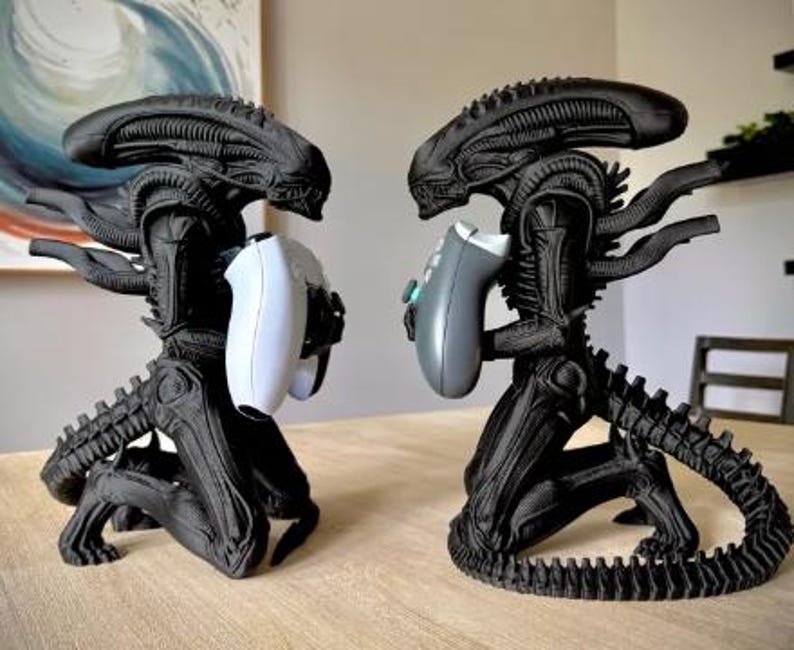 Op de afbeelding: Twee zwarte Xenomorph-figuren in een knielende houding, elk met een videogamecontroller in de hand. Het linkerfiguur houdt een witte controller vast, terwijl het rechterfiguur een grijze controller vasthoudt. Beide figuren hebben gedetailleerde ribbels.