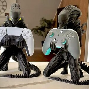 Op de afbeelding: Twee zwarte Xenomorph-figuren in een knielende houding, elk met een videogamecontroller in de hand. Het linkerfiguur houdt een wit-zwarte PlayStation 5-controller vast, terwijl het rechterfiguur een grijze en groenblauwe Xbox-controller vasthoudt.