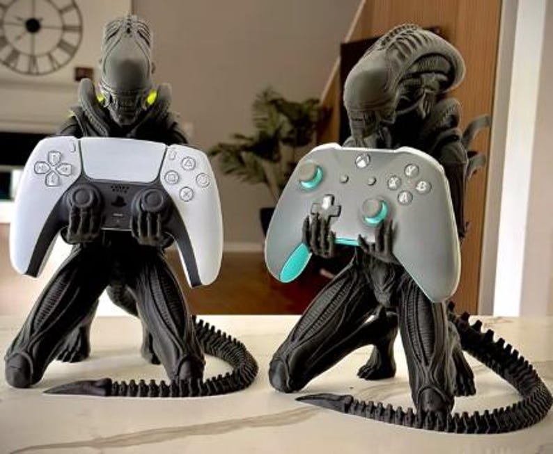 Op de afbeelding: Twee zwarte Xenomorph-figuren in een knielende houding, elk met een videogamecontroller in de hand. Het linkerfiguur houdt een wit-zwarte PlayStation 5-controller vast, terwijl het rechterfiguur een grijze en groenblauwe Xbox-controller vasthoudt.