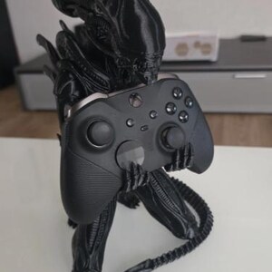 Op de afbeelding: Een zwart 3D-geprint Xenomorph-figuur in een gehurkte houding, die een zwarte draadloze Xbox-controller in de handen houdt. Het wezen heeft een gladde, langwerpige kop, gesegmenteerde ledematen en een lange, puntige staart die naar buiten steekt.
