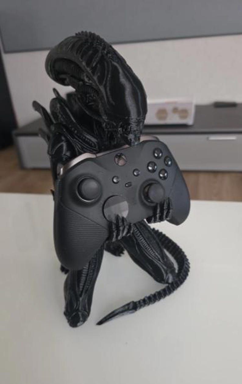 Op de afbeelding: Een zwart 3D-geprint Xenomorph-figuur in een gehurkte houding, die een zwarte draadloze Xbox-controller in de handen houdt. Het wezen heeft een gladde, langwerpige kop, gesegmenteerde ledematen en een lange, puntige staart die naar buiten steekt.