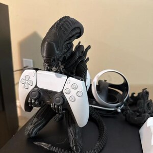 Op de afbeelding: Een zwart Xenomorph-figuur dat dient als controllerstandaard en een wit met zwarte draadloze gamecontroller vasthoudt. Het figuur heeft een lange, gesegmenteerde staart en een gedetailleerd hoofd. Daarnaast staat een wit virtual reality-headsetonderdeel.