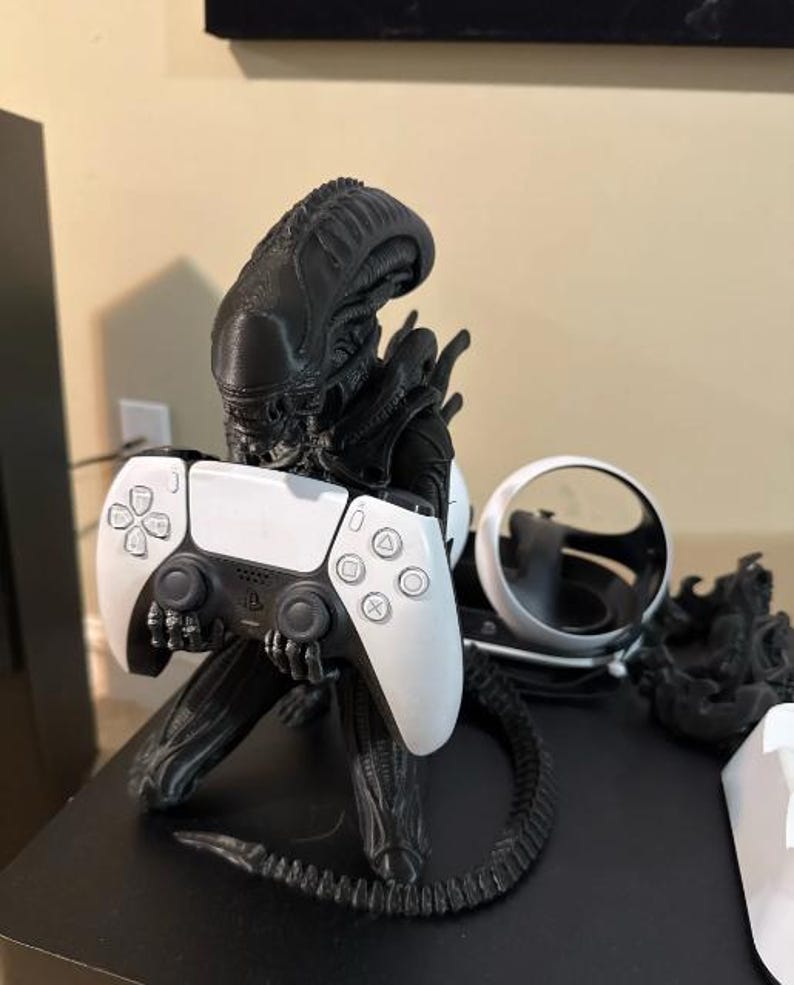 Op de afbeelding: Een zwart Xenomorph-figuur dat dient als controllerstandaard en een wit met zwarte draadloze gamecontroller vasthoudt. Het figuur heeft een lange, gesegmenteerde staart en een gedetailleerd hoofd. Daarnaast staat een wit virtual reality-headsetonderdeel.