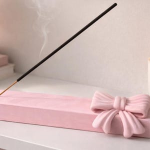Puede incluir: Un porta incienso rosa con un lazo decorativo. Un palito de incienso encendido emite humo. Una vela encendida y libros están en el fondo. El soporte es rectangular y tiene un agujero.