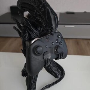 Op de afbeelding: Een zwart 3D-geprint Xenomorph-figuur in een gehurkte houding, met een langwerpig hoofd en een gesegmenteerde staart. Het figuur houdt een zwarte draadloze videogamecontroller vast met twee analoge sticks en een directionele pad.