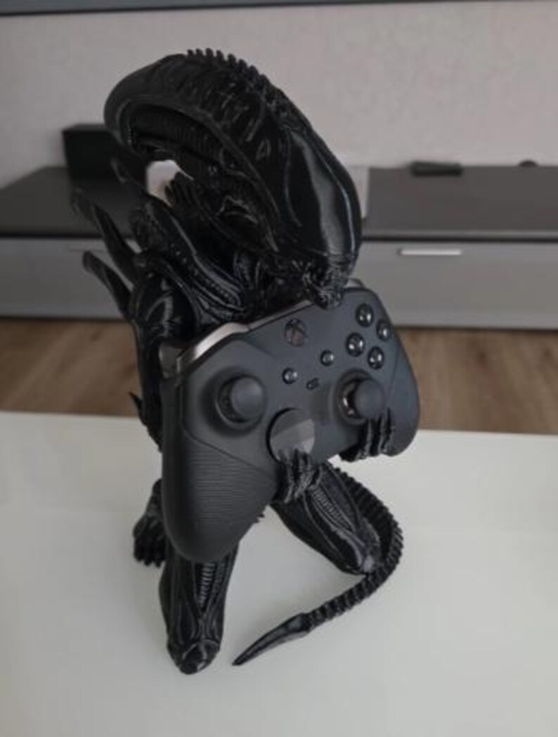 Op de afbeelding: Een zwart 3D-geprint Xenomorph-figuur in een gehurkte houding, met een langwerpig hoofd en een gesegmenteerde staart. Het figuur houdt een zwarte draadloze videogamecontroller vast met twee analoge sticks en een directionele pad.