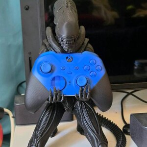 Op de afbeelding: Een zwart Xenomorph-figuurtje in een gehurkte houding, dat een felblauwe draadloze Xbox-controller vasthoudt. Het figuurtje heeft een lange, gesegmenteerde staart die over het oppervlak uitsteekt en een gedetailleerd, langwerpig hoofd met zichtbare tanden.