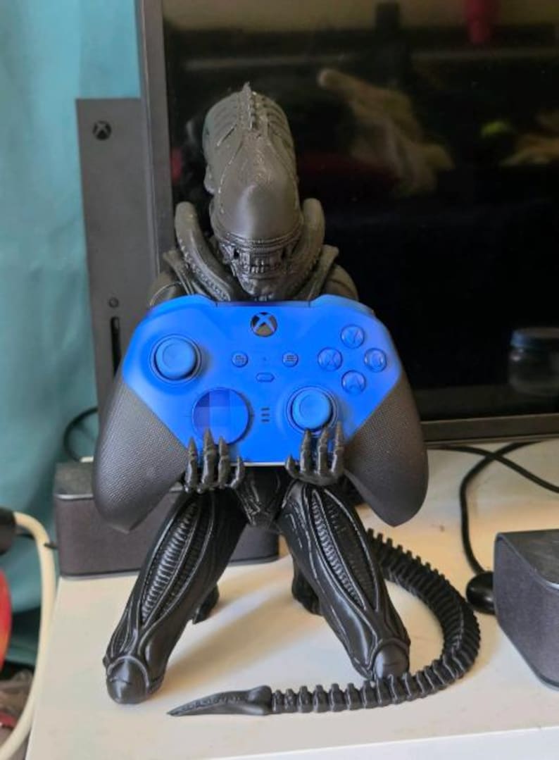 Op de afbeelding: Een zwart Xenomorph-figuurtje in een gehurkte houding, dat een felblauwe draadloze Xbox-controller vasthoudt. Het figuurtje heeft een lange, gesegmenteerde staart die over het oppervlak uitsteekt en een gedetailleerd, langwerpig hoofd met zichtbare tanden.