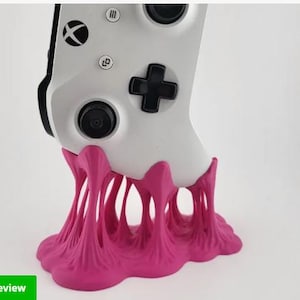 Puede incluir: Un mando de Xbox blanco es sostenido por un soporte rosa vibrante. El soporte tiene un diseño texturizado único que se asemeja a material derretido. El mando presenta un panel direccional y botones negros. La imagen incluye una etiqueta "3D Preview".