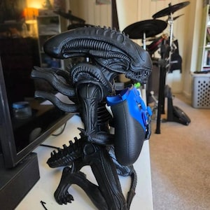 Op de afbeelding: Een zwart Xenomorph-figuur in een knielende houding, met een gladde, langwerpige kop en een geribbelde lichaamsstructuur. Het figuur houdt een blauwe en zwarte videogamecontroller in de handen, met de armen naar voren gericht.