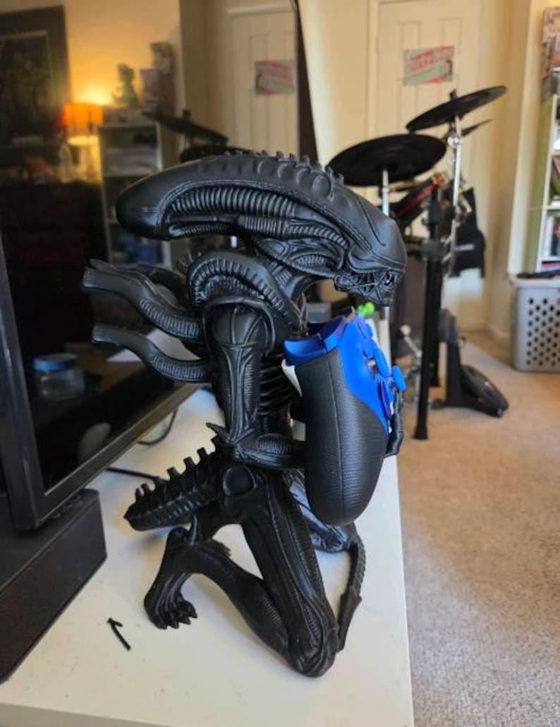 Op de afbeelding: Een zwart Xenomorph-figuur in een knielende houding, met een gladde, langwerpige kop en een geribbelde lichaamsstructuur. Het figuur houdt een blauwe en zwarte videogamecontroller in de handen, met de armen naar voren gericht.