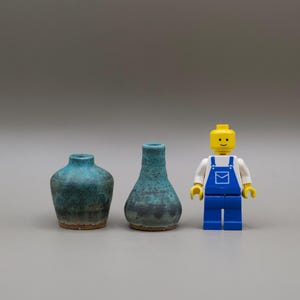 May include: Two miniature ceramic vases with a mottled teal and dark blue glaze, featuring a rounded base and a narrow neck, positioned next to a yellow plastic toy figure wearing blue overalls and a white shirt for scale comparison.
