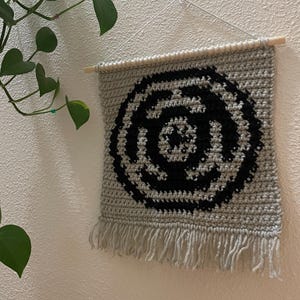 May include: A gray crochet wall hanging with a black circular design. The handmade piece features a fringe at the bottom and is suspended from a wooden dowel. The wall decor is displayed against a white wall with a green plant in the corner.