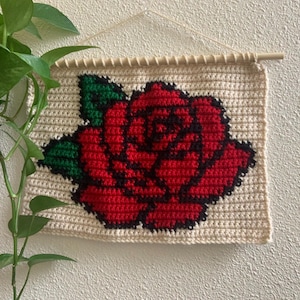 Puede incluir: Tapiz de pared tejido a ganchillo con una rosa roja, hojas verdes y contornos negros sobre un fondo crema. El tapiz está sujeto a un palo de madera para colgarlo.