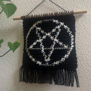 May include: Black crochet wall hanging with a white pentagram design. It has a fringe at the bottom and is suspended from a wooden dowel with black string. The background is a light-coloured wall.