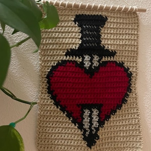 Może przedstawiać: Beżowy szydełkowy obraz na ścianę przedstawiający czerwone serce przebite czarnym i srebrnym sztyletem. Serce jest obrysowane na czarno. Obraz jest przymocowany do drewnianego kołka beżowym sznurkiem.