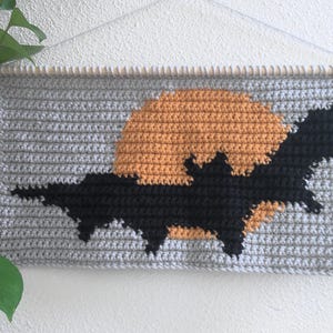 May include: Hand-crocheted wall hanging with a black bat silhouette against an orange moon on a grey background. The piece is mounted on a wooden dowel, ideal for Halloween or spooky decor.