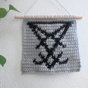 May include: A gray crocheted wall hanging with a black symbol design. The wall hanging is rectangular and is suspended from a wooden dowel. The symbol is a stylized inverted cross with additional elements.