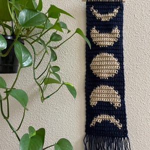 May include: A navy blue crochet wall hanging featuring phases of the moon in a tan color. The wall hanging is rectangular with a fringe at the bottom and a wooden dowel at the top for hanging. A green plant is in the background.
