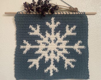 Op de afbeelding: Een handgemaakte, blauw-witte gehaakte wandhanger met een sneeuwvlokontwerp. Gemonteerd op een houten stok en versierd met gedroogde bloemen, creëert het een gezellige, winterse decoratie voor in huis.