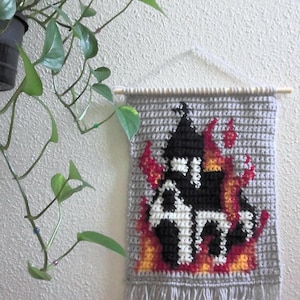 May include: Crocheted wall hanging featuring a black and white house engulfed in flames, with red and yellow accents. The gray background has a fringe at the bottom and is suspended from a wooden dowel.