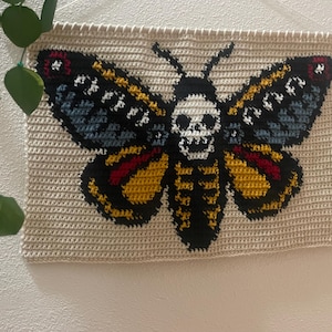 May include: Crocheted wall hanging with a moth design and a skull motif. The moth's wings are black, yellow, red, and blue. The background is a light beige, and the piece is mounted on a wooden dowel. The wall hanging is a decorative item.