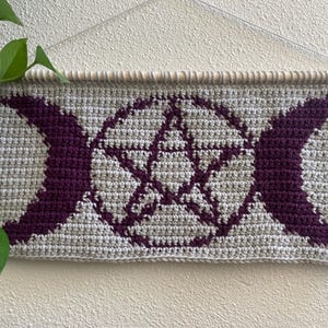 Pode incluir: Tapeçaria de parede de crochê com fundo cinza, design central de pentagrama e luas crescentes roxas escuras. Montada em um bastão de madeira com fio de cor neutra. Uma planta verde é visível no canto superior esquerdo.