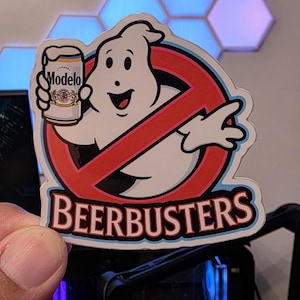 May include: A sticker featuring the Ghostbusters logo with a cartoon ghost holding a Modelo beer can. The text "BEERBUSTERS" is below the logo in a stylized font. The sticker is held up in front of a computer.