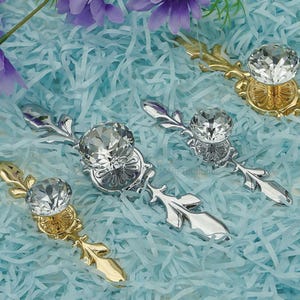 May include: Four decorative cabinet pulls with crystal-like knobs and ornate, leaf-shaped bases. Two are gold-toned, and two are silver-toned. The pulls are arranged on a light blue, textured surface with purple flowers.