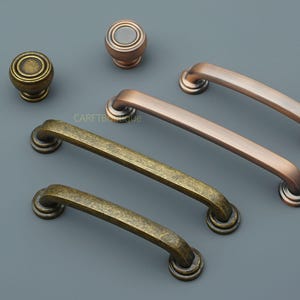 May include: A collection of antique-style cabinet hardware, including knobs and pull handles. The hardware features a variety of finishes, including bronze and copper tones. The pieces are arranged on a gray surface.