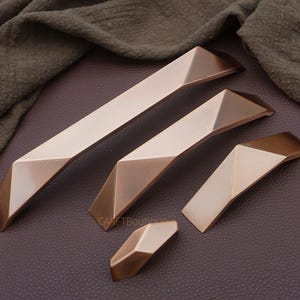 May include: A set of geometric bronze cabinet pulls and knobs. The hardware features a modern design with angled facets and a two-tone finish. The pulls vary in size, with the longest measuring approximately 12 inches. The hardware is displayed on a dark brown surface.